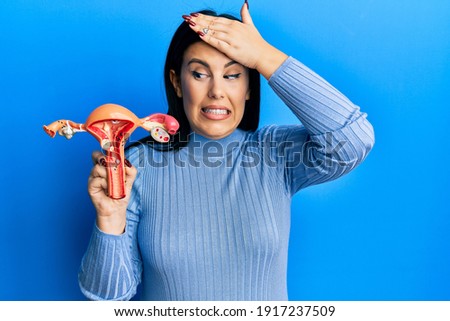 Beautiful brunette woman holding anatomical model of female genital organ stressed and frustrated with hand on head, surprised and angry face  Royalty-Free Stock Photo #1917237509