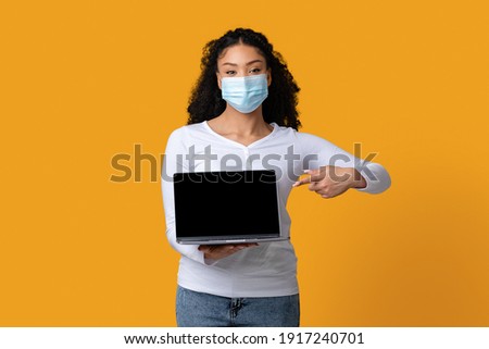 Young black woman wearing protective medical mask holding and pointing at laptop with black screen, standing over yellow studio background and looking at camera, mockup image with copy space