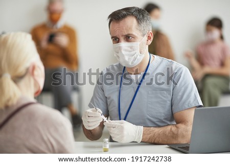 Portrait of mature male doctor wearing mask while consulting patients waiting for covid vaccination in clinic