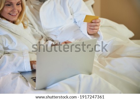 Copy-space photo of a smiley woman using laptop and staying in bed with her boyfriend holding credit card