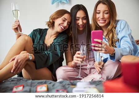Front view of smiling ladies with champagne flutes looking through the selfies on the cellphone