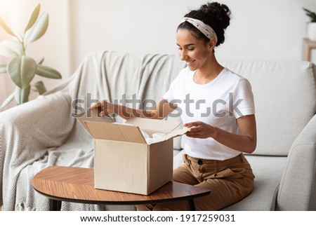 Happy young woman unpacking delivery box, sitting on couch by tea table, home interior. Satisfied customer cheerful lady opening package, ordering clothes or cosmetics on Internet, copy space Royalty-Free Stock Photo #1917259031
