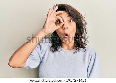 Young hispanic woman wearing casual winter sweater doing ok gesture shocked with surprised face, eye looking through fingers. unbelieving expression. 
