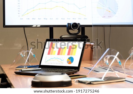 Laptop and television display with chart ,diagram presentation and face shield on table in meeting room with clear acrylic sheet separates the center
