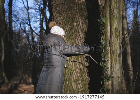 Waist-up portrait of warmly dressed woman hugging huge tree trunk in the woods. 