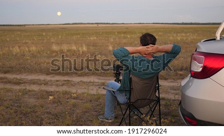 A man traveler in car stopped at a camping in evening and takes pictures with a camera next to car. Freelance photographer working outdoors, beautiful moments for photos and videos. Driver is resting