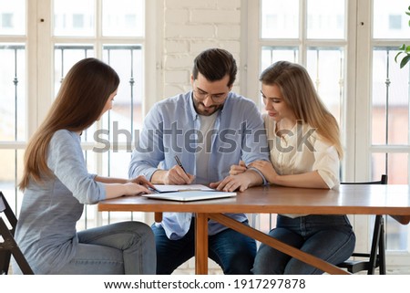 Happy young clients signing paper contract with professional broker or realtor, after discussing agreement details. Confident saleswoman watching millennial couple putting signature, purchasing house.