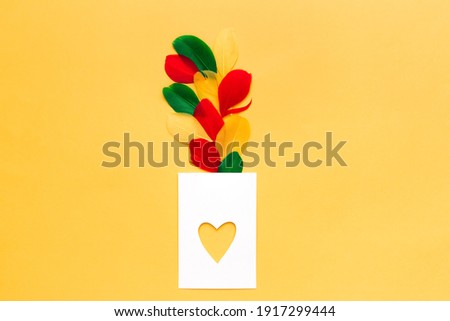 Blank white card in shape of heart, on yellow background, with colored feathers, top view. Greeting card template. Love concept. Copy space.
