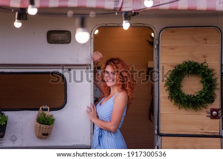 A beautiful slender girl with red curly hair and a gorgeous smile in a blue sundress sits in the doorway of her trailer. A tender mother travels with her happy family in a caravan car. Budget vacation Royalty-Free Stock Photo #1917300536