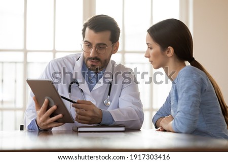 Focused young male doctor pointing at digital computer tablet screen, sharing health tests laboratory results to serious female patient or explaining medical insurance benefits at meeting in clinic. Royalty-Free Stock Photo #1917303416