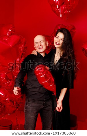 Bald man and young woman in black stand with heart-shaped balloons on red background
