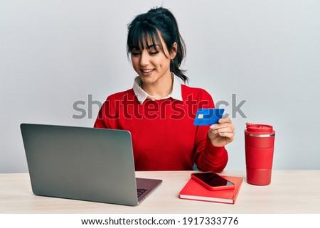 Young brunette woman with bangs working at the office using laptop and credit card looking positive and happy standing and smiling with a confident smile showing teeth 