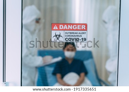 Closeup of label with notice of danger with covid-19 outbreak message on door having a young female patient with doctors dressed in PPE kit making notes of progress