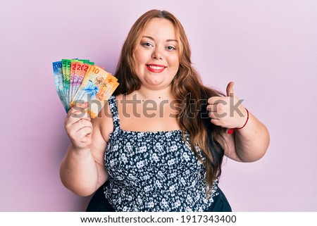 Plus size caucasian young woman holding swiss franc banknotes smiling happy and positive, thumb up doing excellent and approval sign 