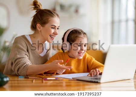 Cheerful little schoolgirl in headphones talking to the teacher while studying remotely via laptop at home with happy mom sitting nearby and giving support Royalty-Free Stock Photo #1917395237