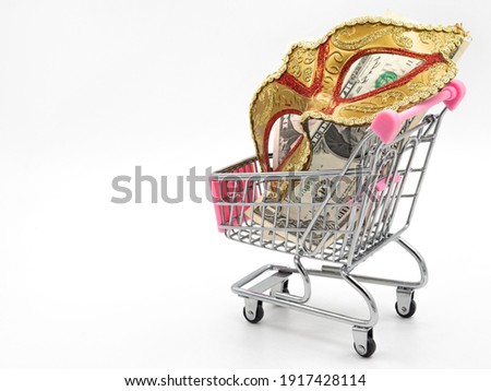 A traditional Venetian golden carnival mask with money (dollar cuppures) in a toy shopping cart on a white background. Concept of preparation for the annual Brazilian festival, costume rental.