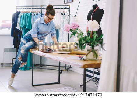 Pensive female fashion designer sitting on table with takeaway hot drink drawing sketches while working on design for new clothes in modern workspace