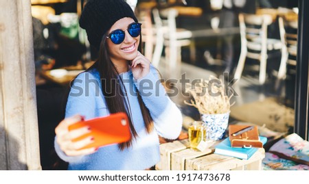 Happy millennial influencer shooting positive video vlog using smartphone front camera during leisure in sidewalk cafe, cheerful hipster girl in trendy sunglasses taking selfie pictures on mobile