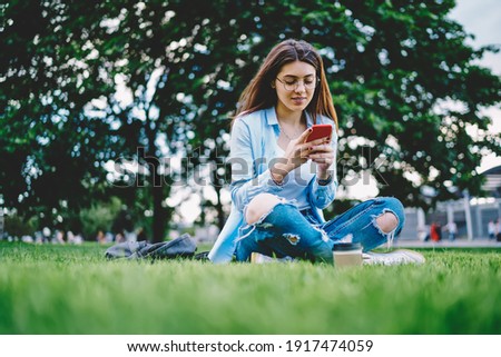 Caucasian hipster girl in classic eyewear using cellphone gadget for online messaging connect to 4g wireless in park, young female blogger networking social media while installing smartphone app