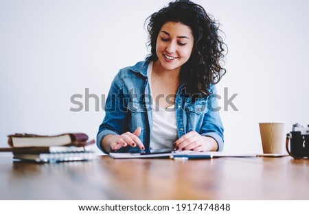 Curly female blogger with cute smile browsing web page on modern touch pad technology using public internet connection in cafe, millennial student searching education information on digital tablet