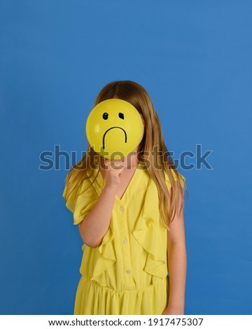 A young child is holding a yellow balloon to her face with a sad face drawing on it with a blue background for a childhood sadness or unhappiness concept.