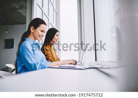 Female colleagues smiling in coworking office cooperating using laptop computers for online browsing, cheerful employees sharing media content during brainstorming programming and networking