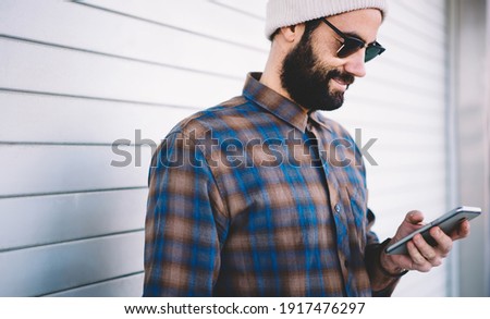Middle Eastern male blogger in stylish sunglasses writing email text for sending via smartphone application, millennial hipster guy browsing touristic information on website connected to 4g internet