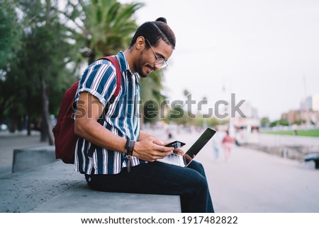 Side view of positive ethnic man in trendy clothes and eyeglasses working remotely on laptop and browsing smartphone on stone parapet on street with palms Royalty-Free Stock Photo #1917482822