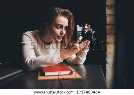 Young female in white jumper shooting with old fashioned analog camera sitting at table with book near window and looking away
