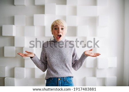 Excited female in casual clothes with short blond hair and opened mouth looking at camera spreading arms standing near wall with geometric decorations