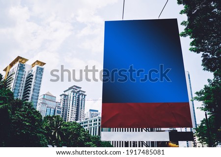 Big blue with red empty billboard surrounded tall modern buildings located in green district of megapolis at daytime with cloudy sky on background