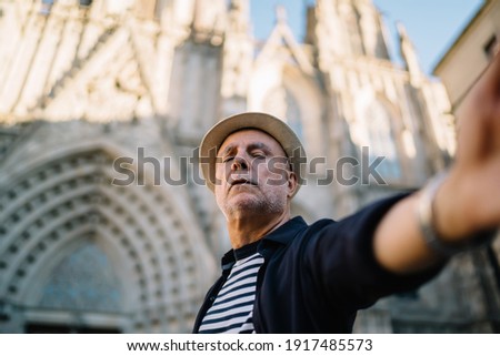 Elderly Caucasian influencer 60 years old shooting travel vlog during solo journey trip for exploring historic city, aged male tourist in straw hat photographing himself during sightseeing