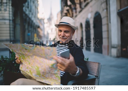 Carefree male tourist reading orientation info on location map holding smartphone technology in hand and resting on city bench, elderly man in straw hat searching destination during journey holidays