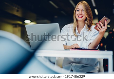 Low angle of smiling adult lady dressed in white shirt looking away while working remotely on computer and using smartphone at table in workplace