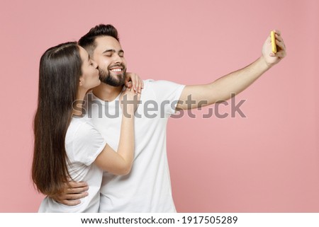 Young cheerful couple two friends bearded man brunette woman in white basic blank print design t-shirts doing selfie shot on mobile cell phone isolated on pastel pink color background studio portrait