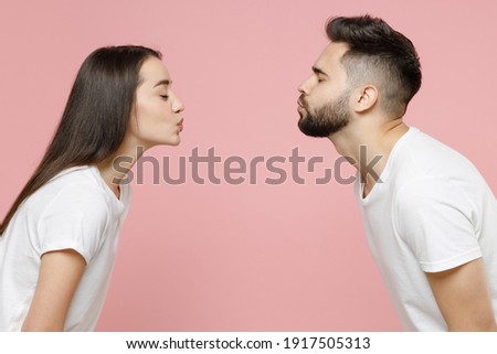 Side profile view young cheerful couple two friends man woman in white basic t-shirts kissing each other with closed eyes while standing face to face isolated on pastel pink background studio portrait Royalty-Free Stock Photo #1917505313