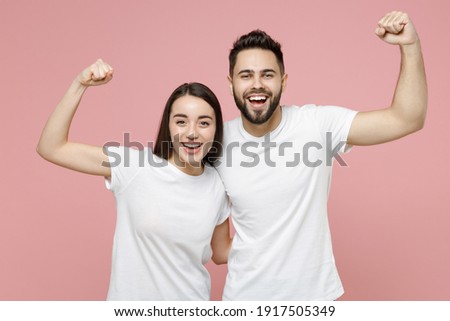 Young cheerful strong sporty fitness couple two friends man woman 20s in white basic blank print design t-shirts showing biceps muscles on hand isolated on pastel pink color background studio portrait Royalty-Free Stock Photo #1917505349