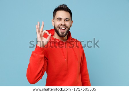 Young caucasian smiling happy bearded attractive handsome positive man 20s wearing casual red orange hoodie show ok okay gesture isolated on blue background studio portrait People lifestyle concept