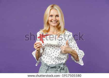 Funny elderly gray-haired blonde woman lady 40s 50s years old in white dotted blouse standing hold in hand gift certificate showing thumb up isolated on bright violet color background studio portrait