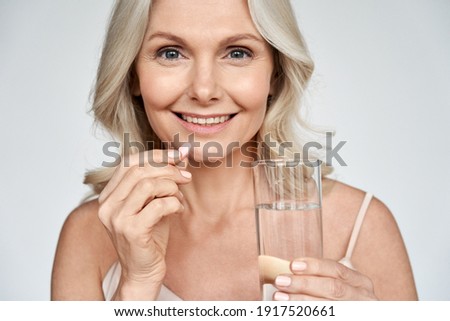Smiling happy healthy middle aged 50s woman holding glass of water taking dietary supplement vitamin pink pill isolated on white background. Old women multivitamins antioxidants for anti age beauty. Royalty-Free Stock Photo #1917520661
