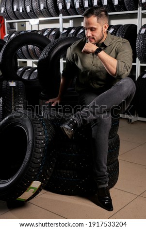 male customer sit thinking which of tires to buy, caucasian guy in casual wear sits in contemplation in auto service shop, surrounded by black tires for car