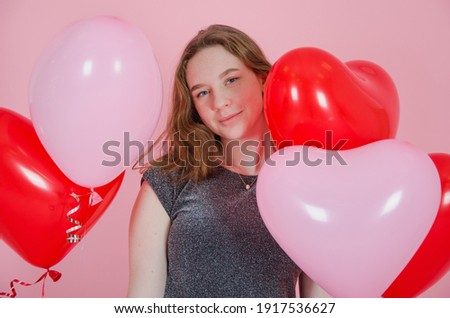 a girl in a gray dress holds heart-shaped balloons on a pink background and laughs
