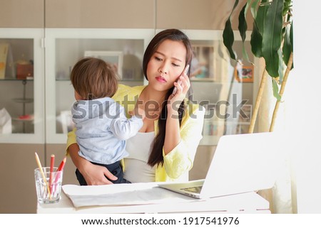 Busy and stressed mother talking on phone with one hand holding her baby while working from home on computer PC. Tired woman using smartphone. Housewife overwork. Social distancing during coronavirus. Royalty-Free Stock Photo #1917541976