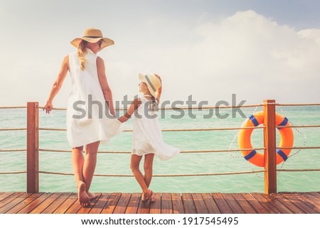 Suntanned woman and girl in white dresses enjoy sea view at the wooden pier. Vacation, get away, travel concept. Toned image, place for text