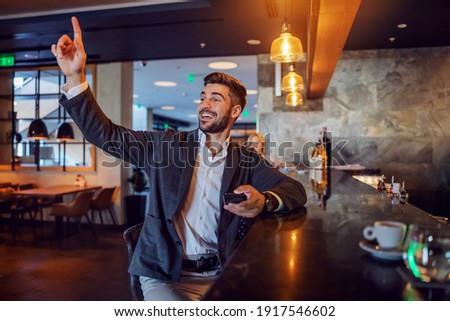 A kind businessman sits at a bar asking for a check. He calls the waiter with one hand while holding the phone in the other. Positive attitude, break time, pause, social media network
