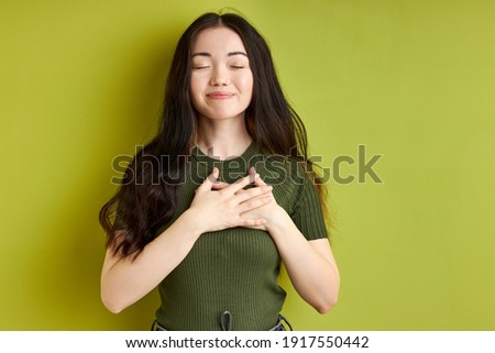 brunette female expressing thanks holding hands on chest with eyes closed, isolated over green studio background. portrait. people emotions concept
