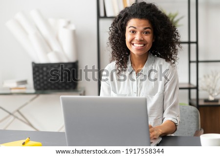 Excited young African American female office worker businesswoman with Afro hairstyle sitting at the desk with laptop, looking at the camera. Portrait of a working mixed-race woman with positive vibe Royalty-Free Stock Photo #1917558344