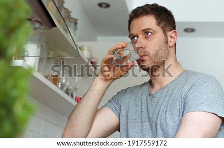 Unshaven man drinks water from a glass in the kitchen.