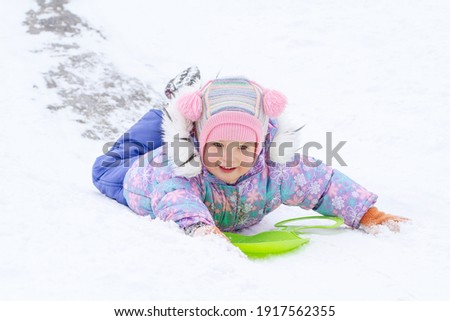A little pretty girl in a purple overalls slides down an ice slide, winter children's games, a child in the park in winter.
