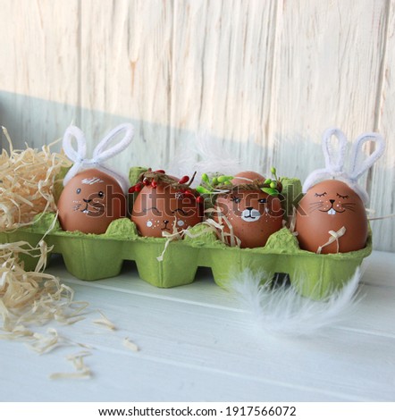 Cute easter eggs with animal faces in a green egg holder on a white wooden background with feathers and straw, do it yourself easter decor
